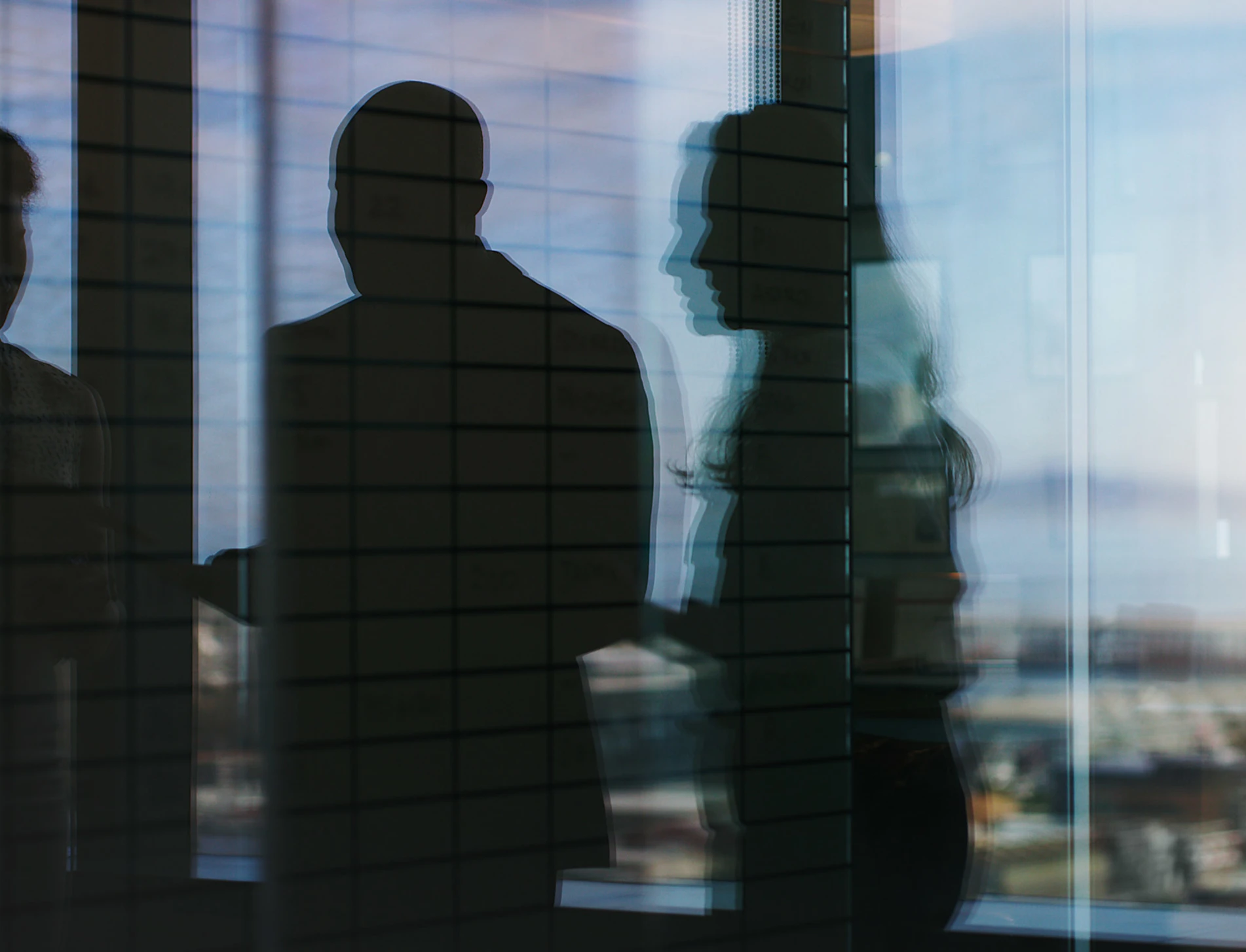 Silhouettes of four people behind a glass panel with a grid pattern, with a blurred city skyline visible beyond.