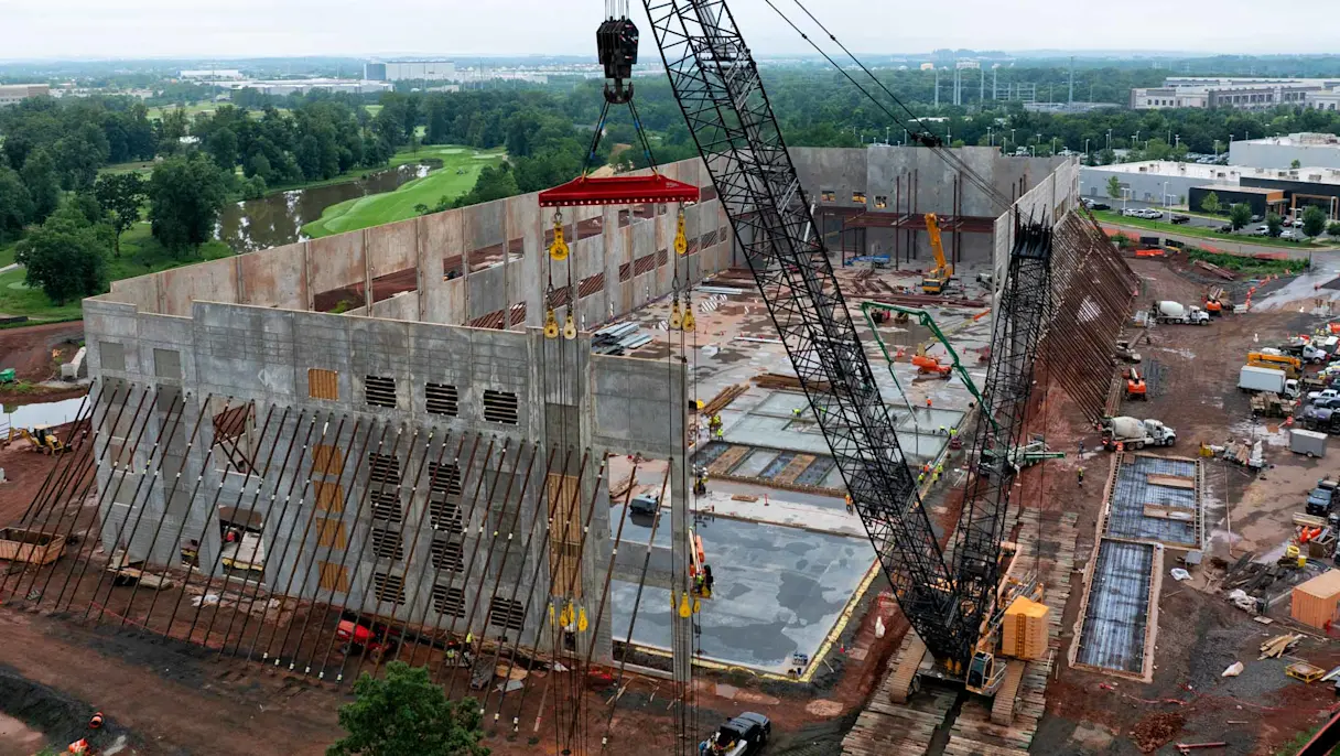 Aerial view of a large construction site with a crane lifting concrete panels, surrounded by roads and buildings.