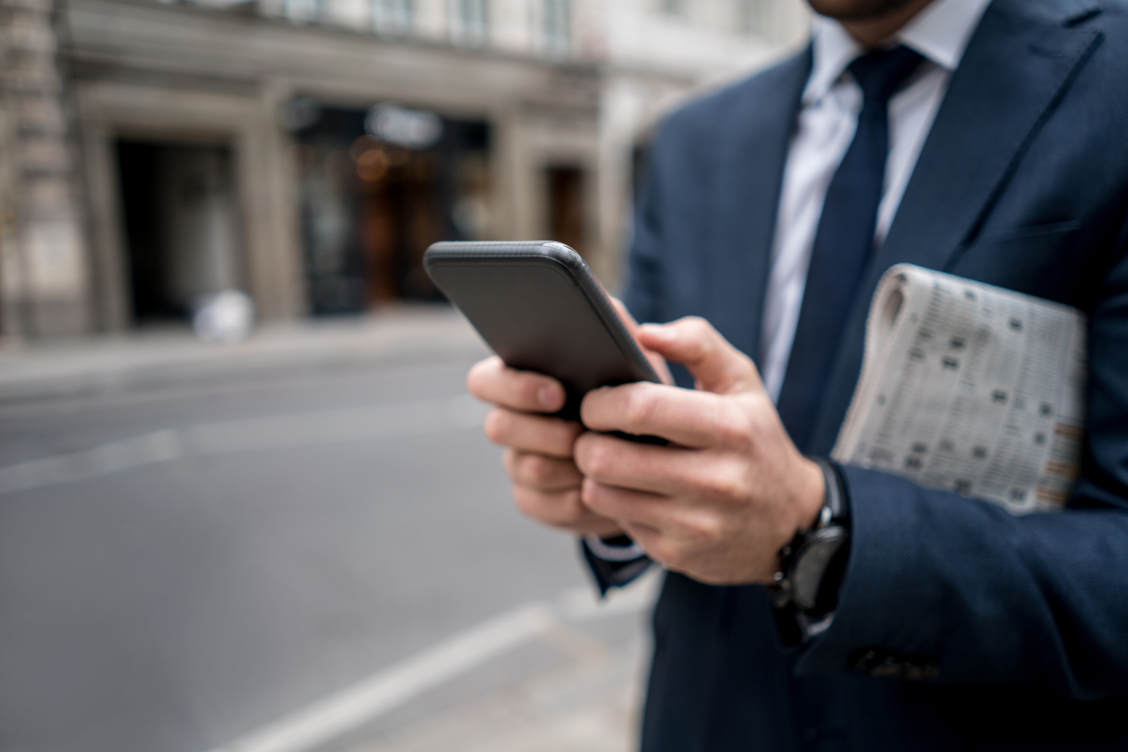Man in a blue suit holding a smartphone and a folded newspaper on a city street, focusing on the phone screen.
