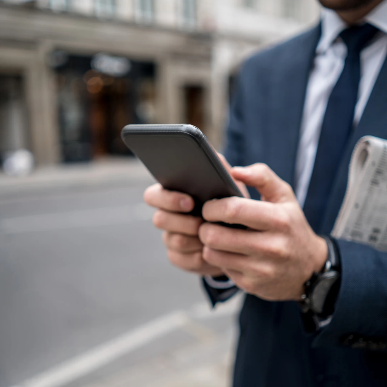 Man in a blue suit holding a smartphone and a folded newspaper on a city street, focusing on the phone screen.