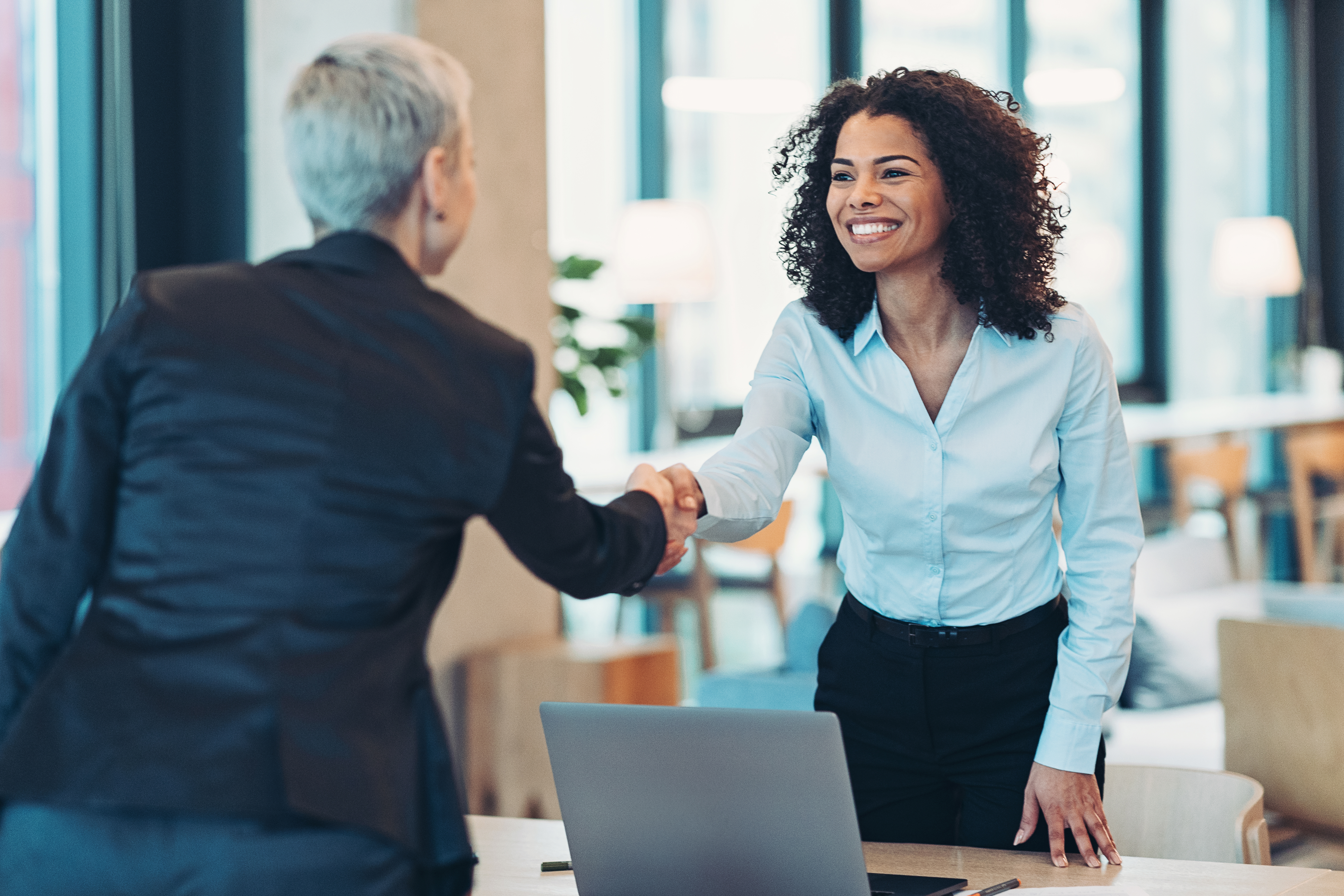 Two businesswomen shaking hands across a desk in a modern office with a laptop on the table.