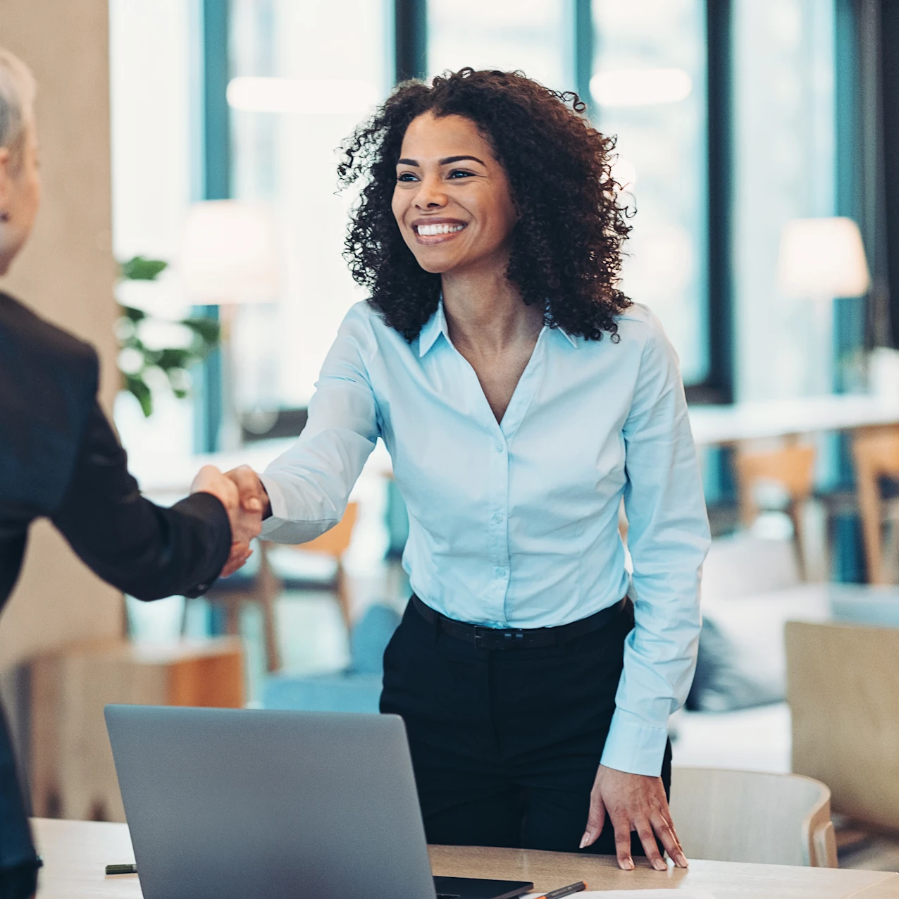Two businesswomen shaking hands across a desk in a modern office with a laptop on the table.