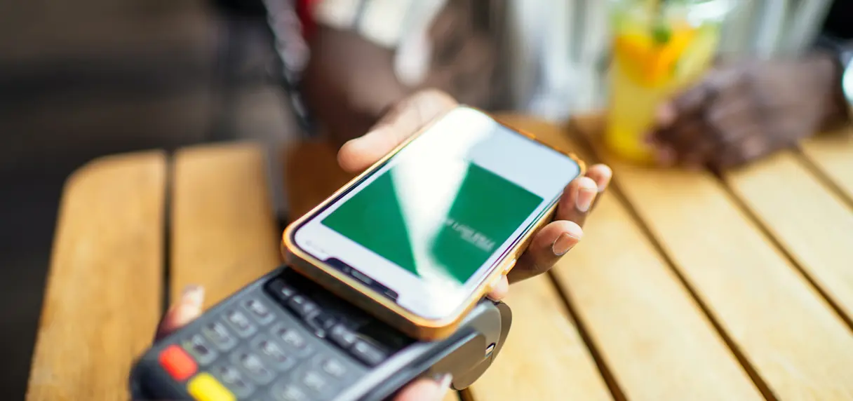 Close-up of a person using a smartphone to make a contactless payment on a card reader over a wooden table with a blurred drink in the background.