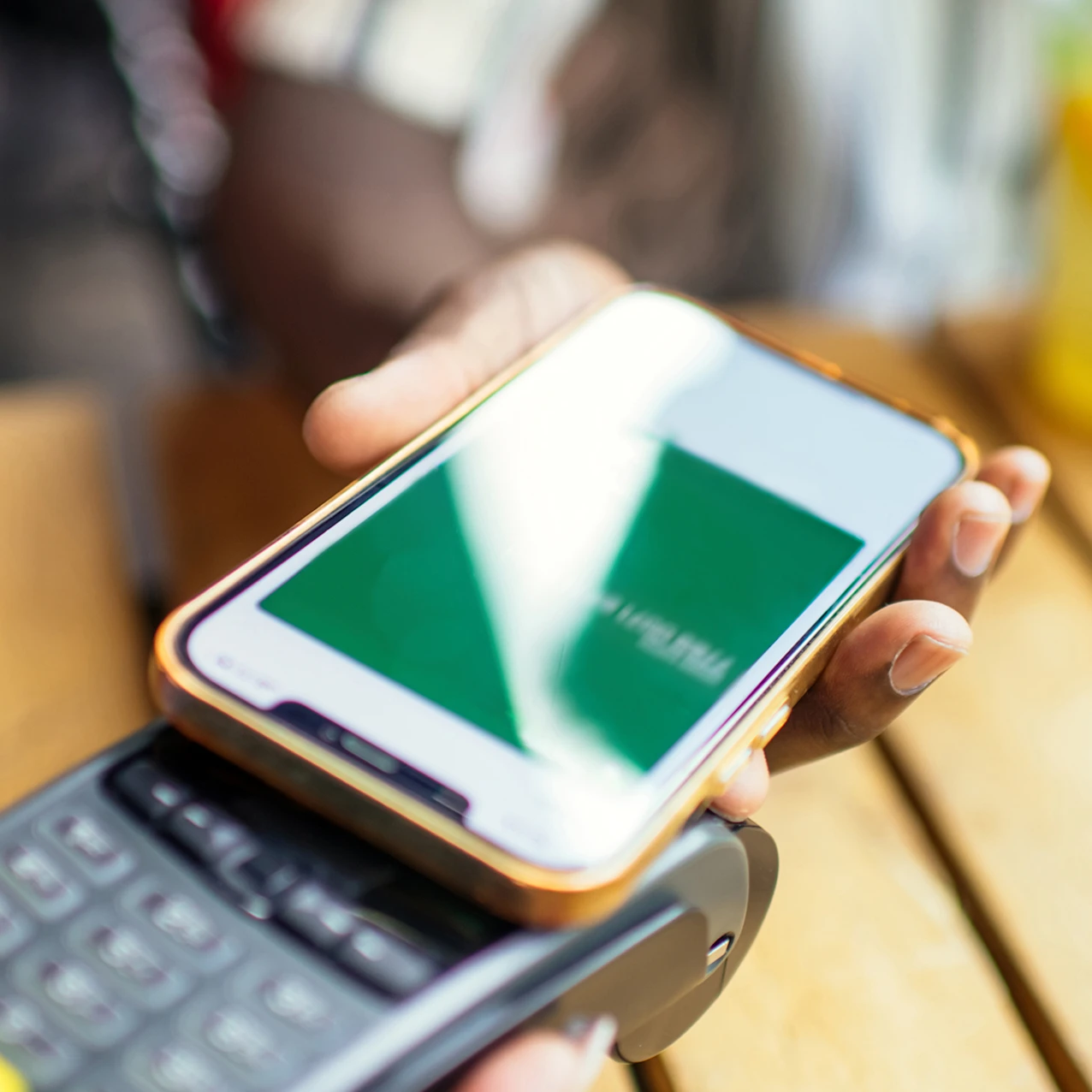 Close-up of a person using a smartphone to make a contactless payment on a card reader over a wooden table with a blurred drink in the background.