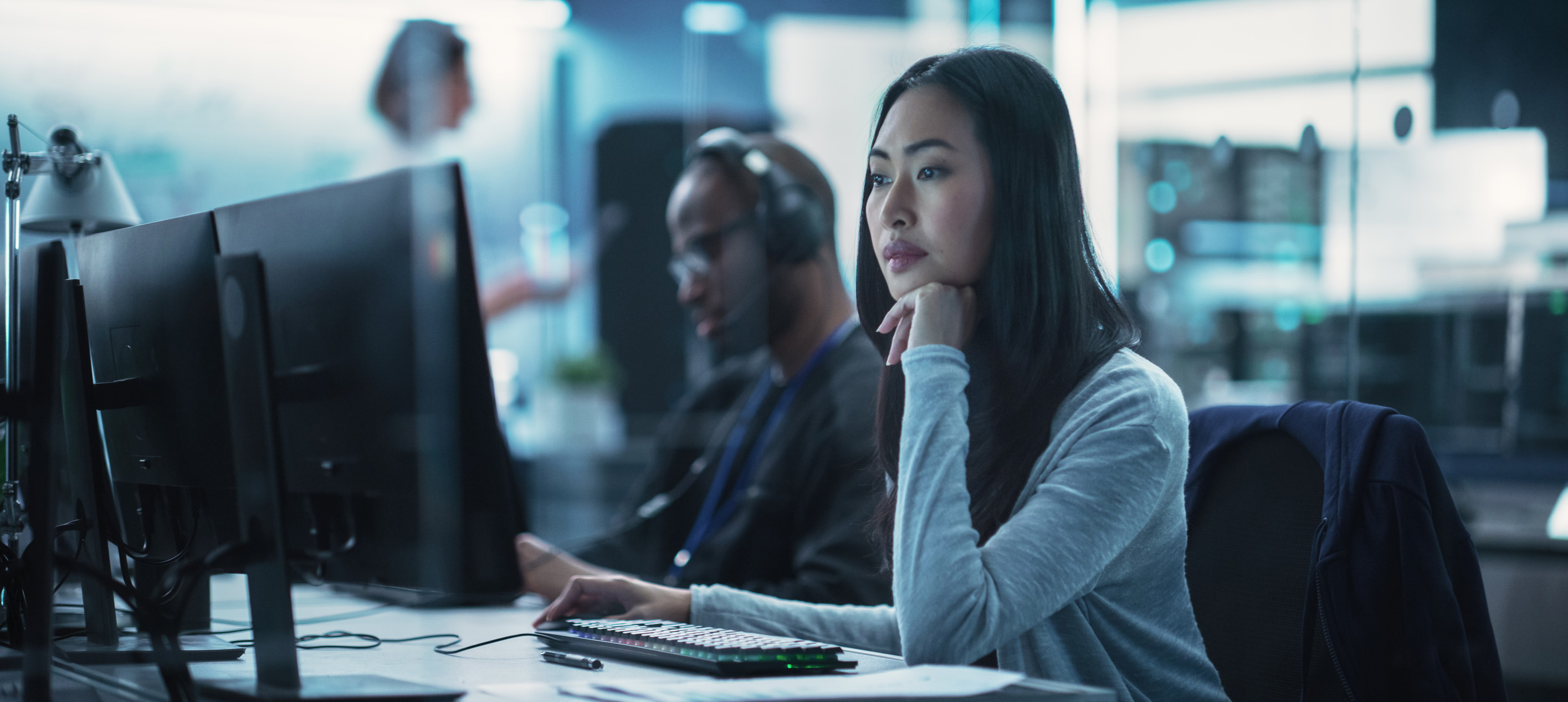 Woman working at a computer workstation in a modern office with colleagues in the background.