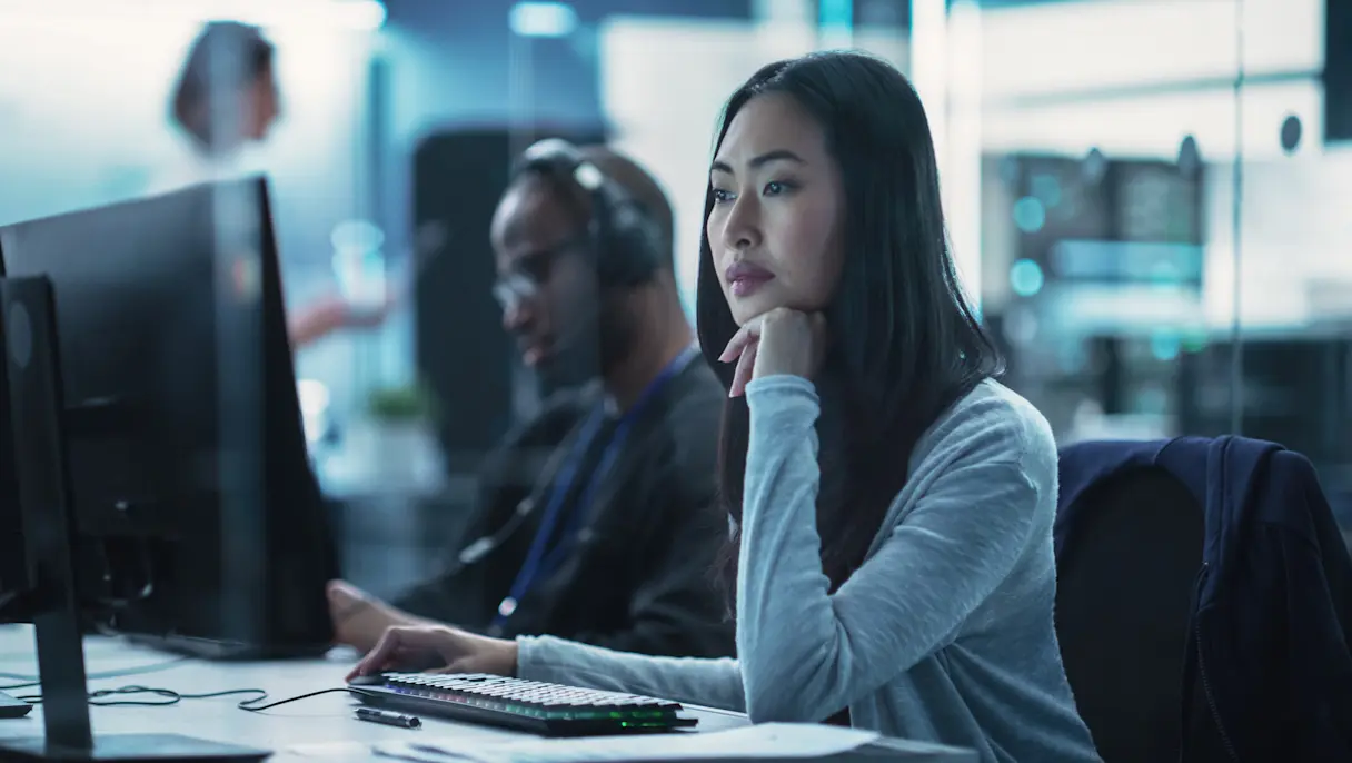 Woman working at a computer workstation in a modern office with colleagues in the background.