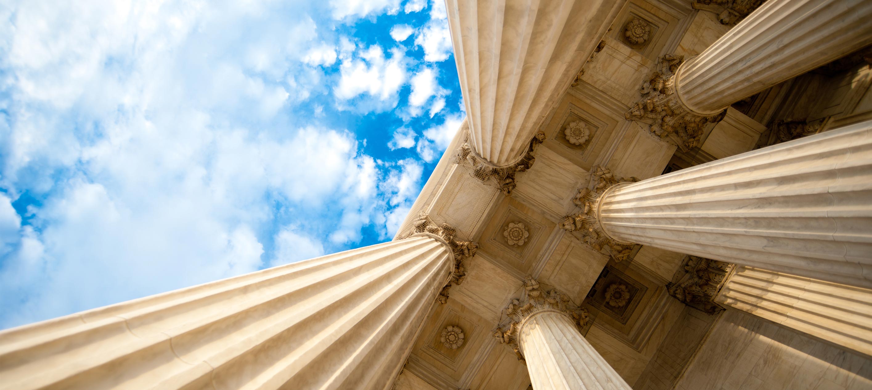 Upward view of classical stone columns and an ornate ceiling on a government building against a blue sky.