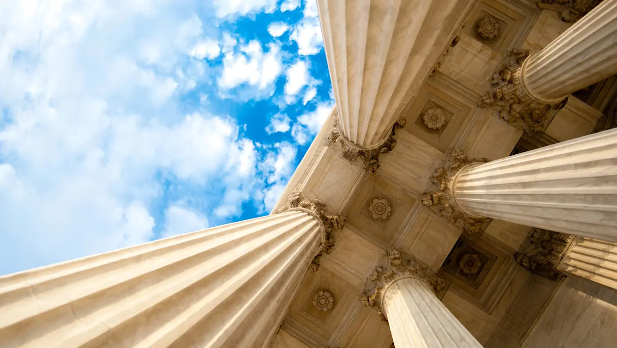 Upward view of classical stone columns and an ornate ceiling on a government building against a blue sky.
