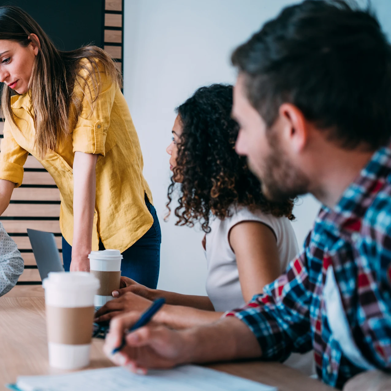 Four colleagues in a meeting room collaborate around a wooden table with laptops and coffee cups, one standing and pointing at a laptop screen.