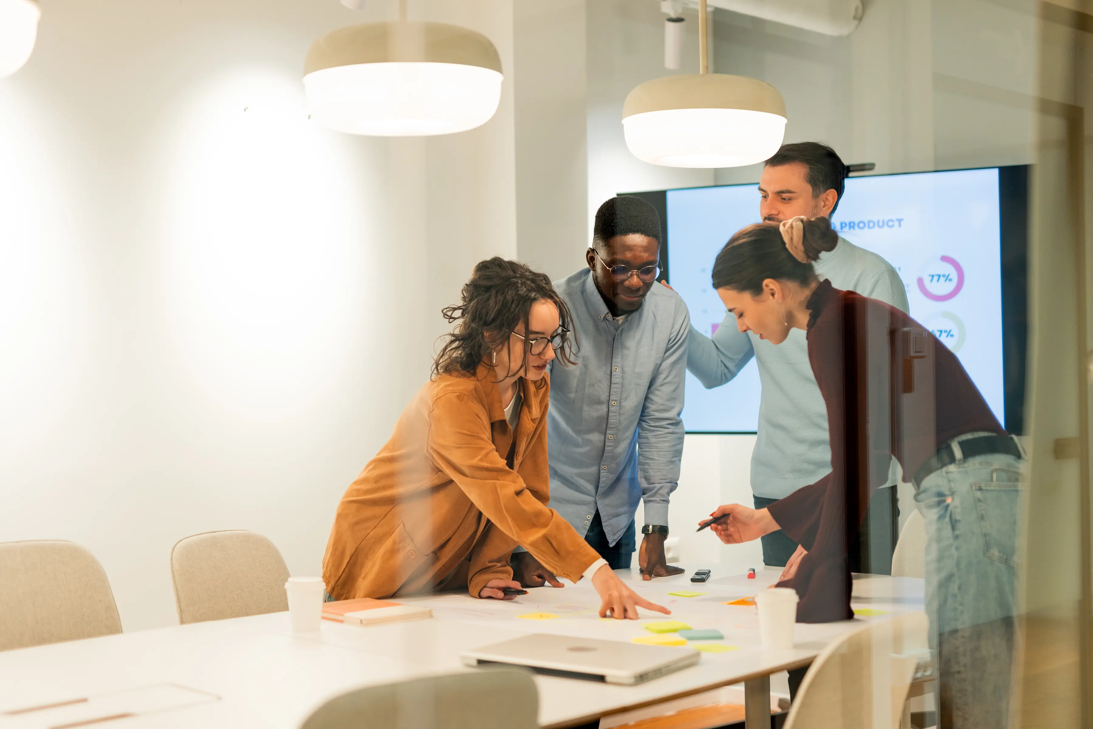 Four colleagues leaning over a conference table with sticky notes and documents, with a data dashboard visible on a screen behind them.