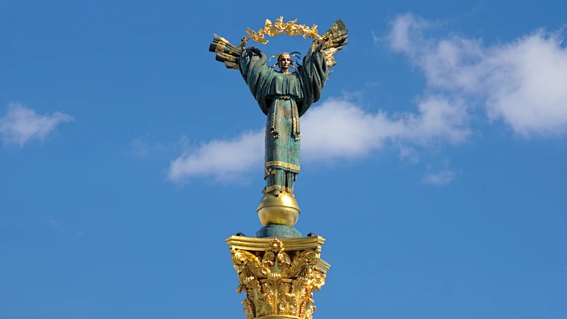 Statue of a robed female figure holding a golden wreath above her head atop a decorative golden column against a blue sky with a few clouds.