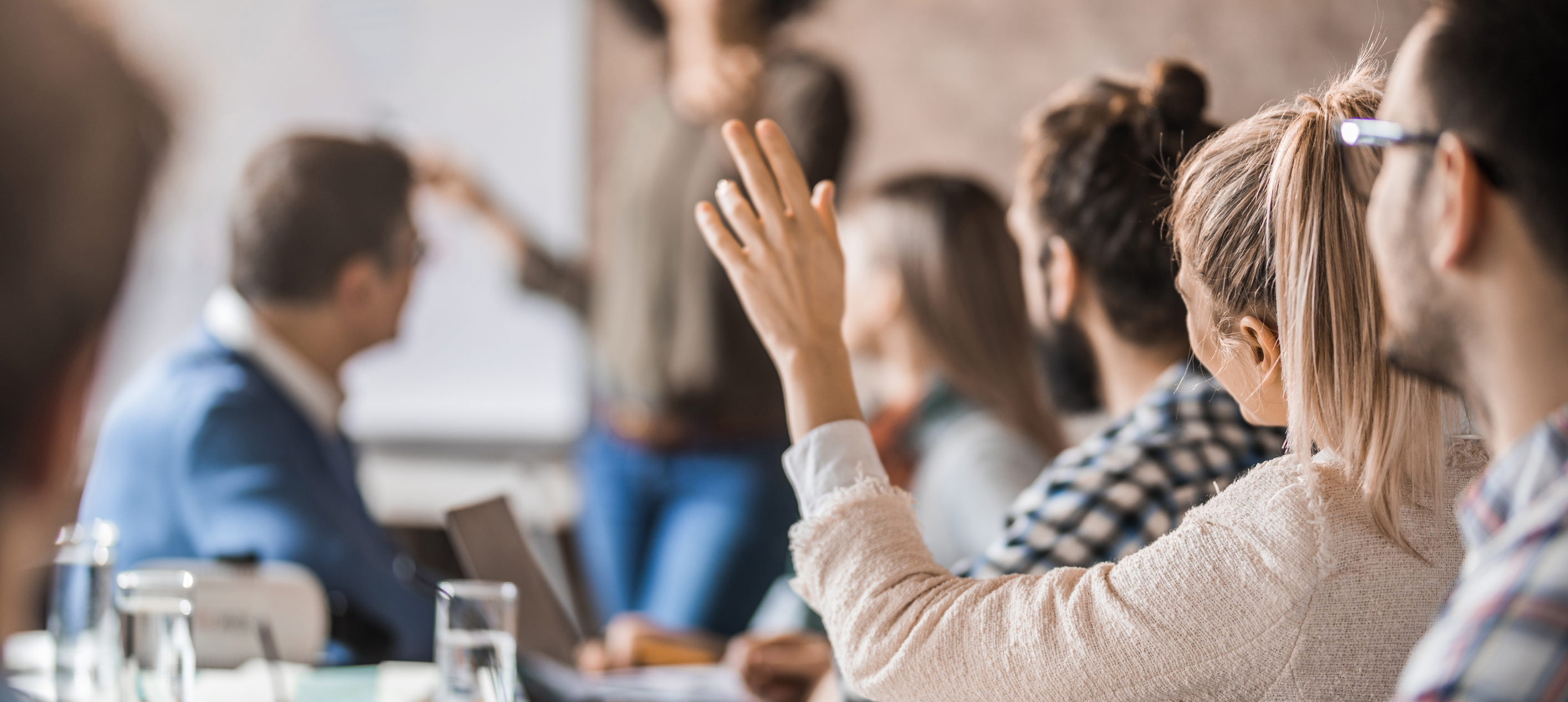 A woman raising her hand in a meeting room while a presenter stands at a whiteboard in the background.