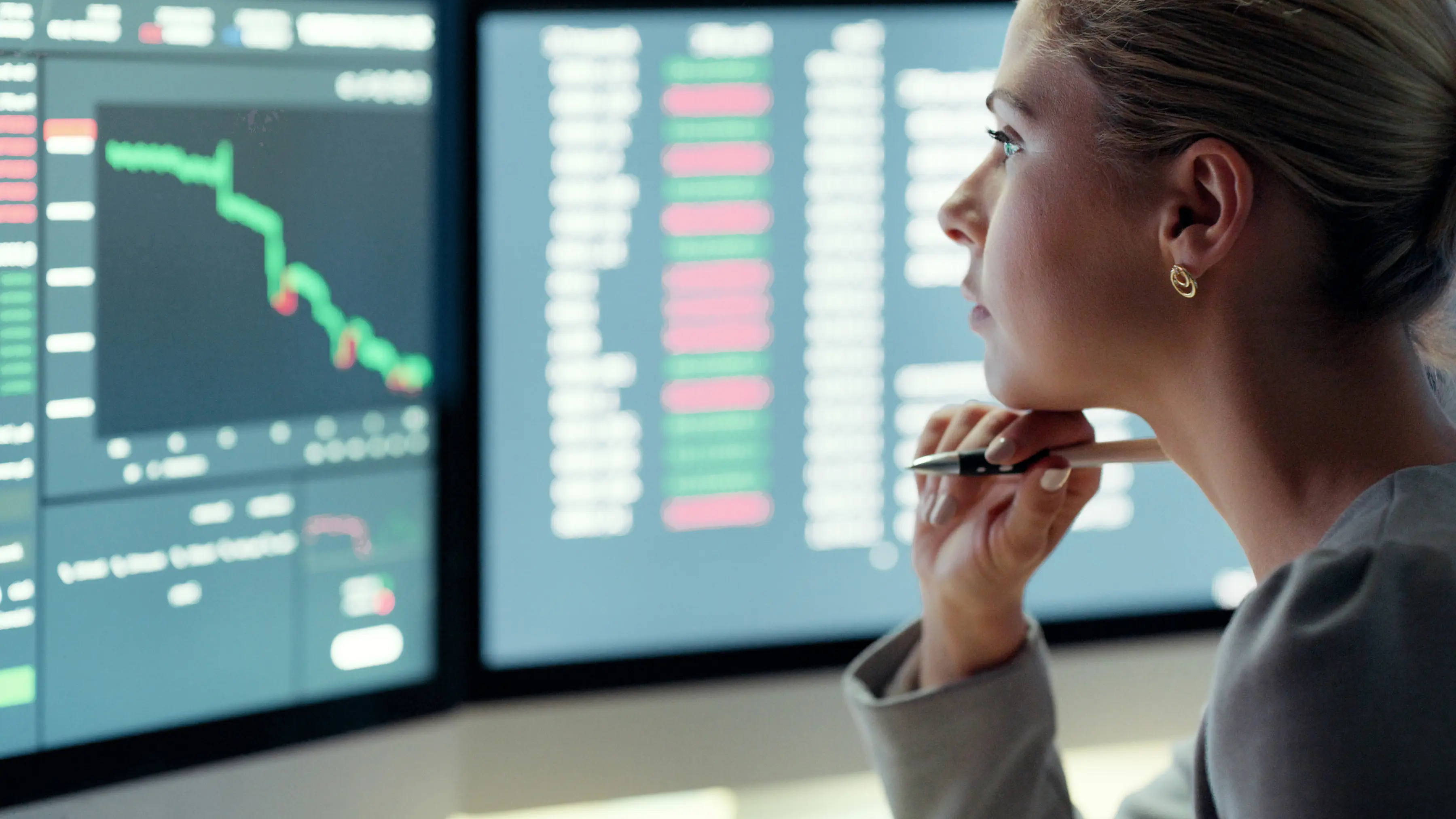Person holding a pen, looking at multiple screens displaying financial charts and data with red and green indicators.