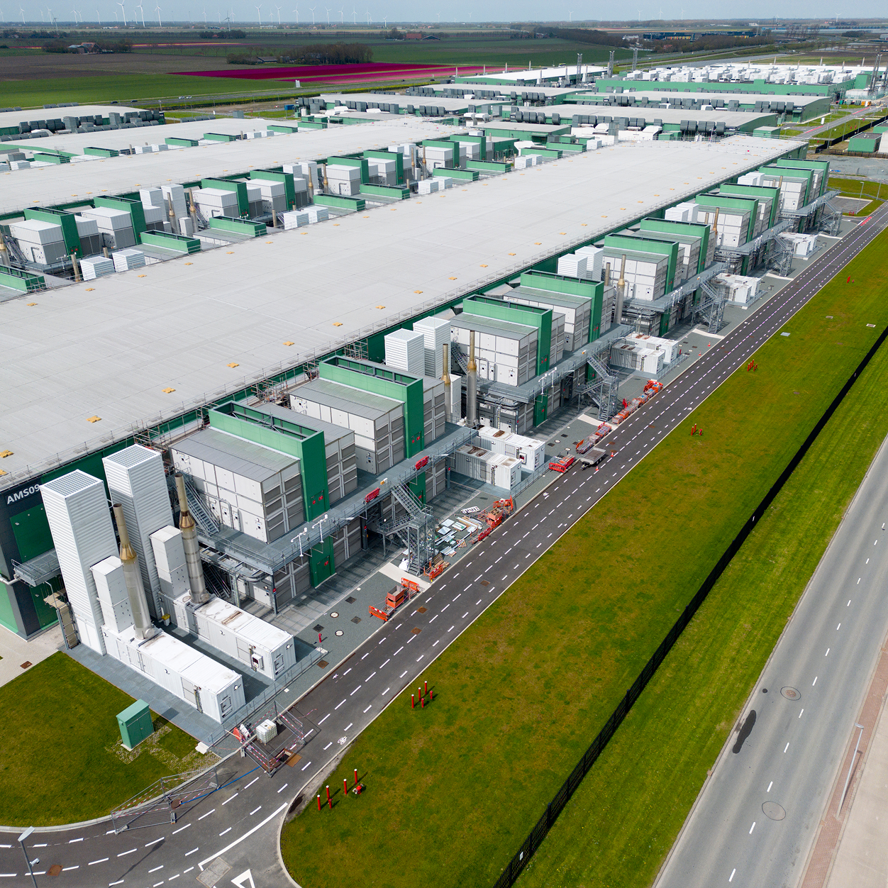 Aerial view of a large data center with rows of green-and-white server buildings next to a road and green fields, with wind turbines on the horizon.