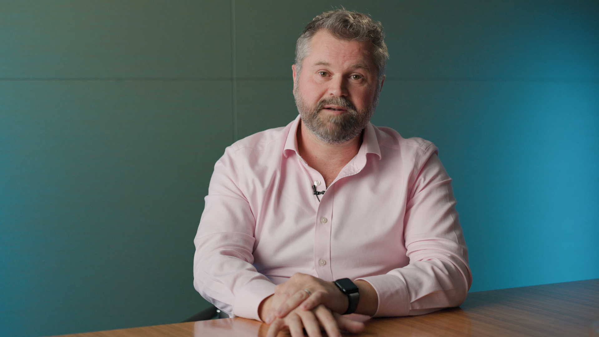 Man wearing a light pink button-up shirt and smartwatch sits at a wooden table with hands folded, against a teal wall background.