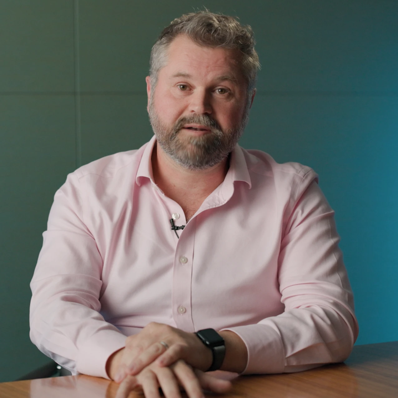 Man wearing a light pink button-up shirt and smartwatch sits at a wooden table with hands folded, against a teal wall background.