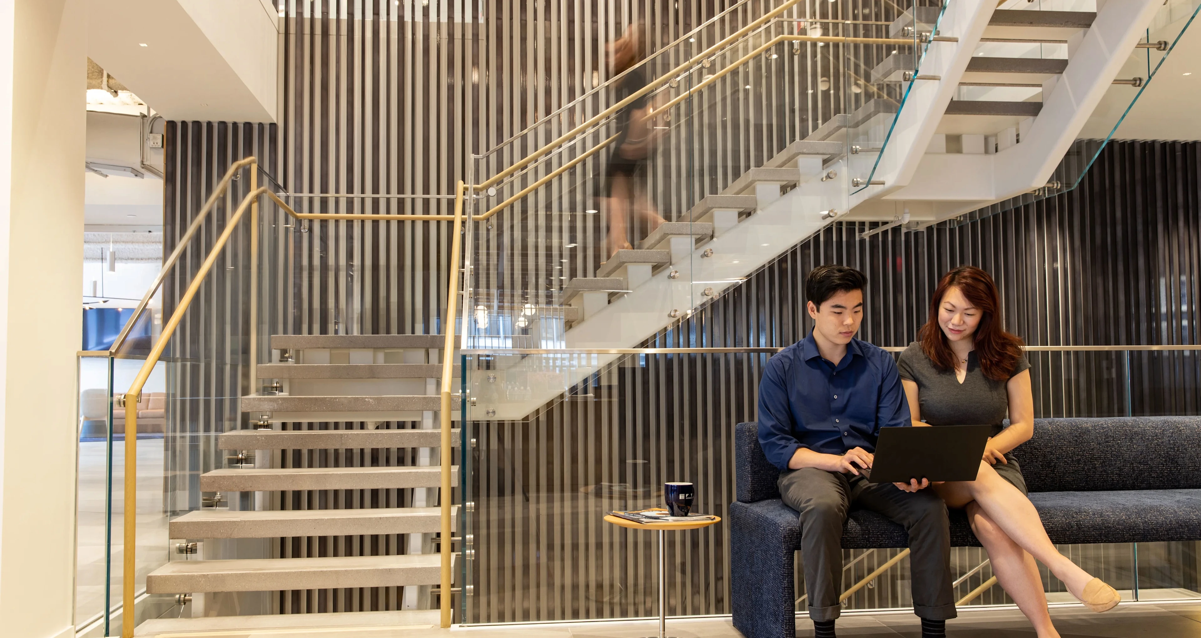 Two young professionals sit on a dark blue sofa in a modern office lobby, collaborating over a laptop near glass stairs with golden handrails.