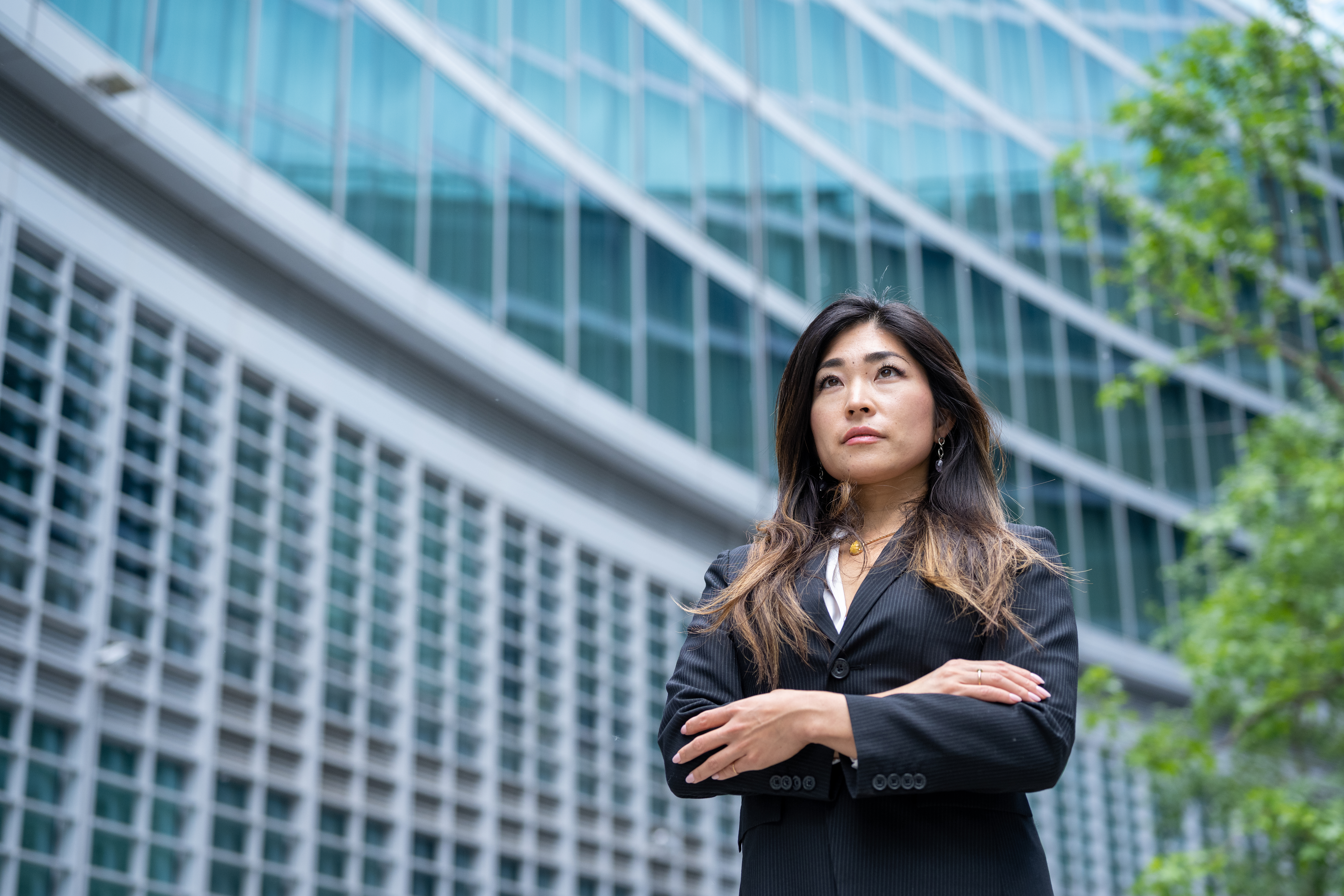 A person wearing a black suit stands confidently with arms crossed in front of a modern glass building, surrounded by greenery.