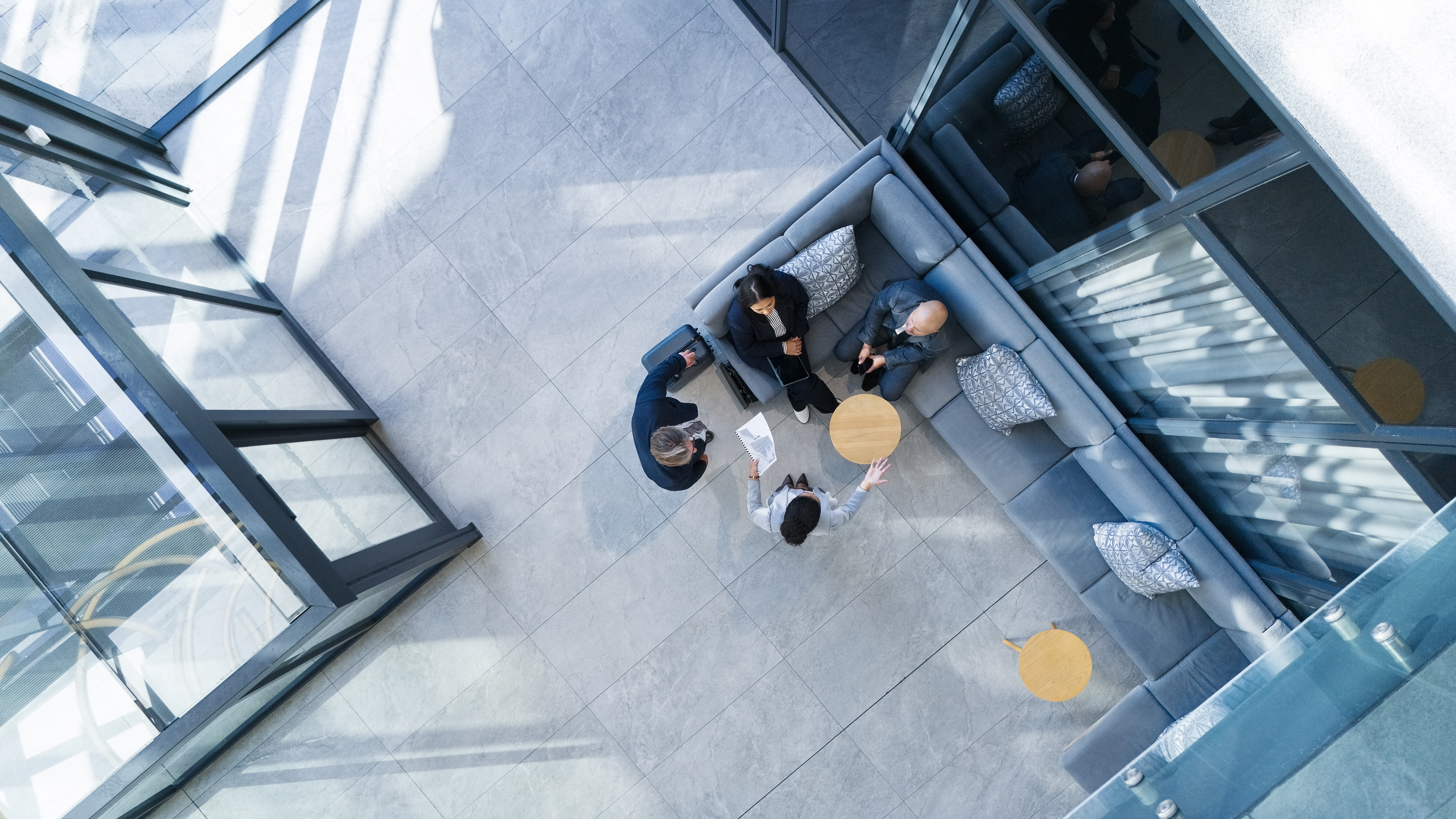 Overhead view of four people having a discussion in a modern office seating area with gray sofas and round wooden tables, surrounded by large glass windows.