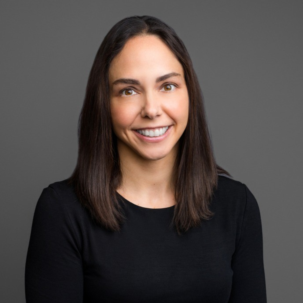 Person with medium-length straight brown hair wearing a black long-sleeve top, photographed against a plain gray background.