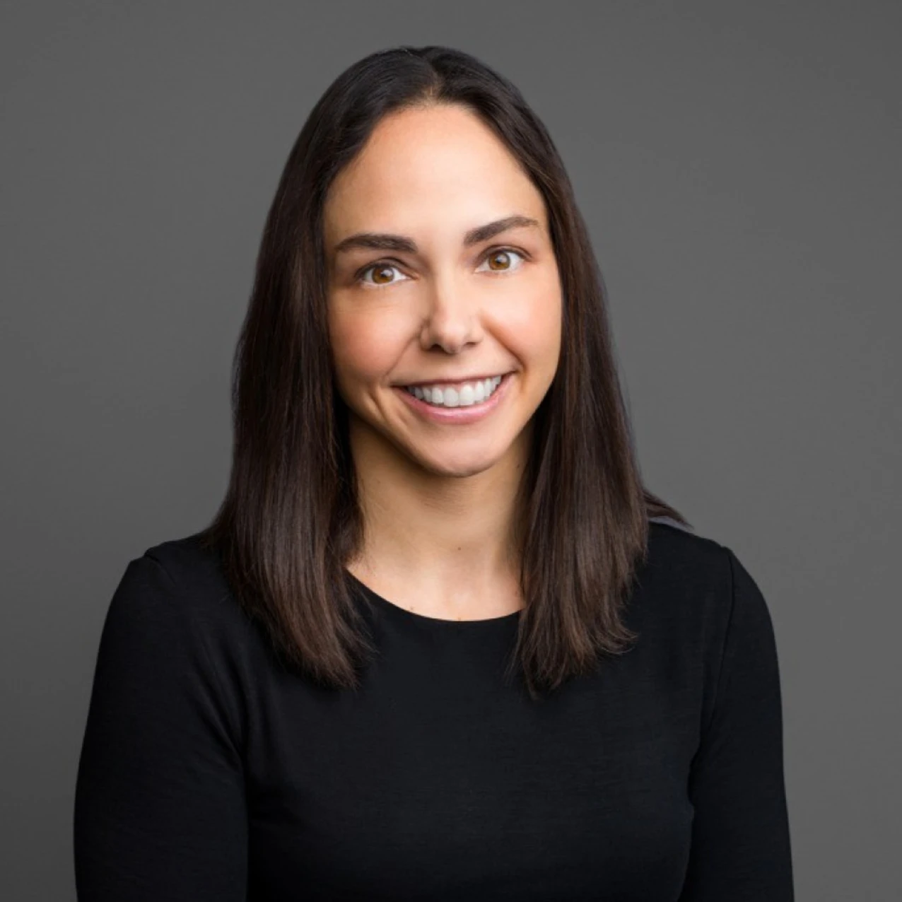 Person with medium-length straight brown hair wearing a black long-sleeve top, photographed against a plain gray background.
