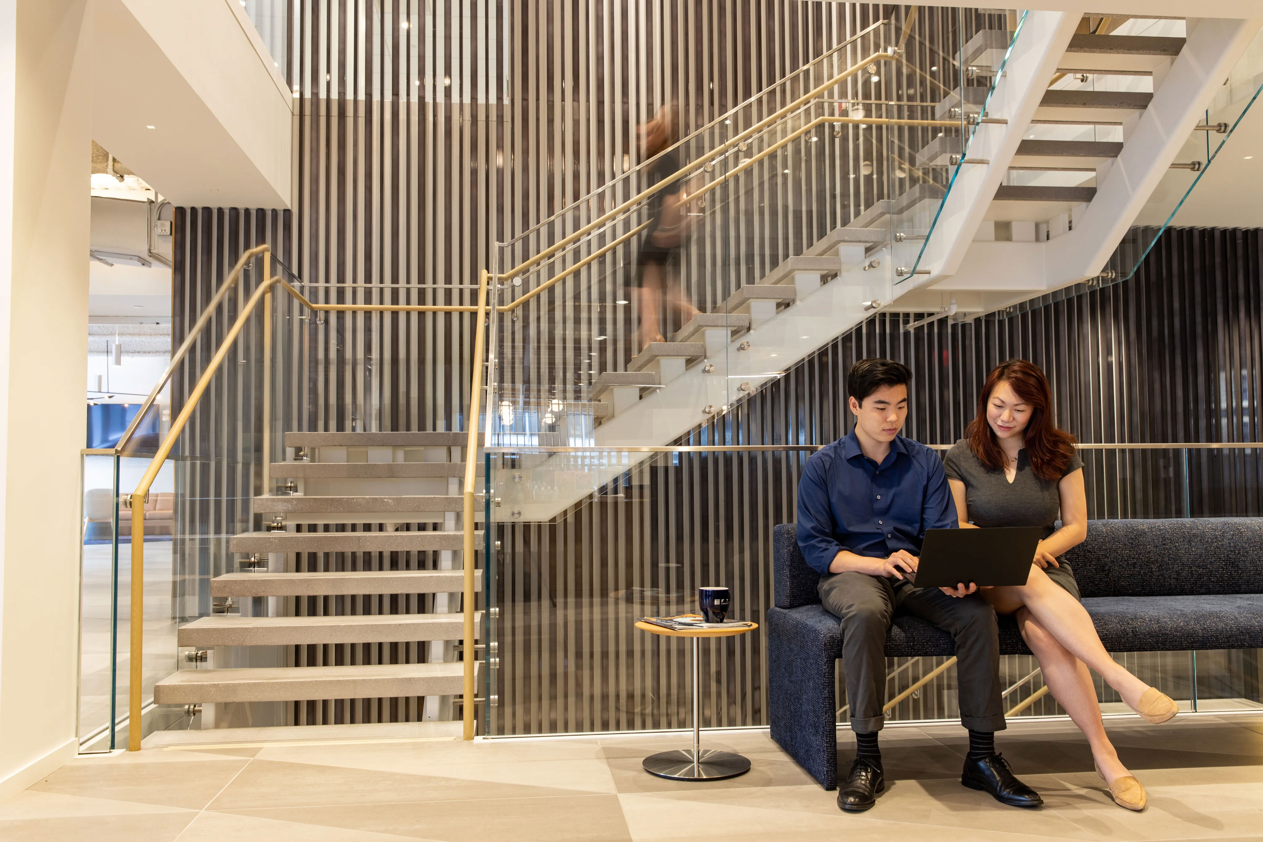 Two colleagues sit on a blue couch in a modern office lobby, looking at a laptop together near a glass staircase with a person walking upstairs blurred in motion.