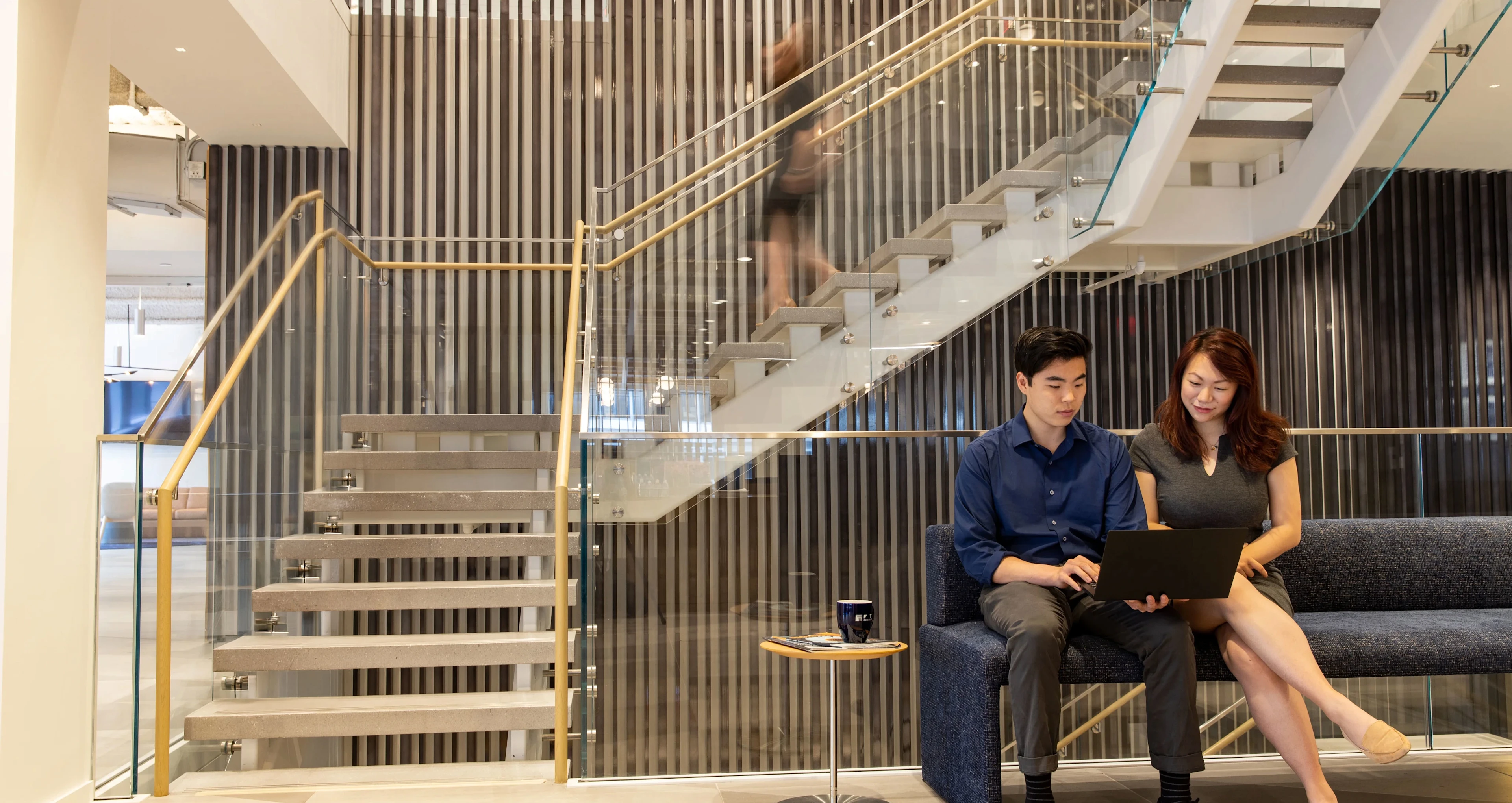 Two colleagues sit on a blue couch in a modern office lobby, looking at a laptop together near a glass staircase with a person walking upstairs blurred in motion.