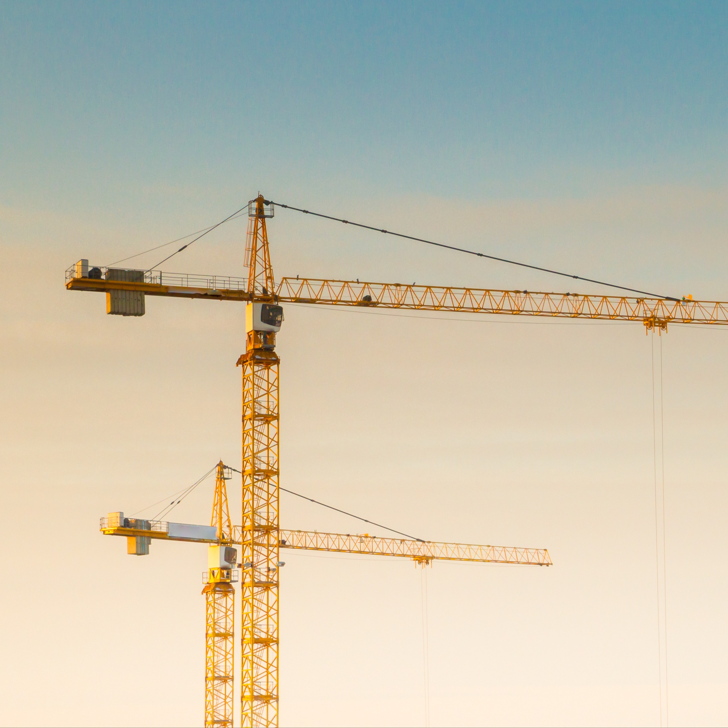 Two yellow construction cranes against a soft gradient sky, with the taller crane in the foreground and the shorter one behind it.