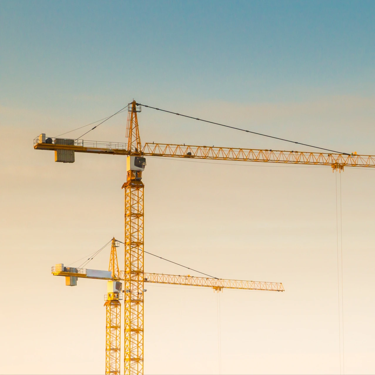 Two yellow construction cranes against a soft gradient sky, with the taller crane in the foreground and the shorter one behind it.
