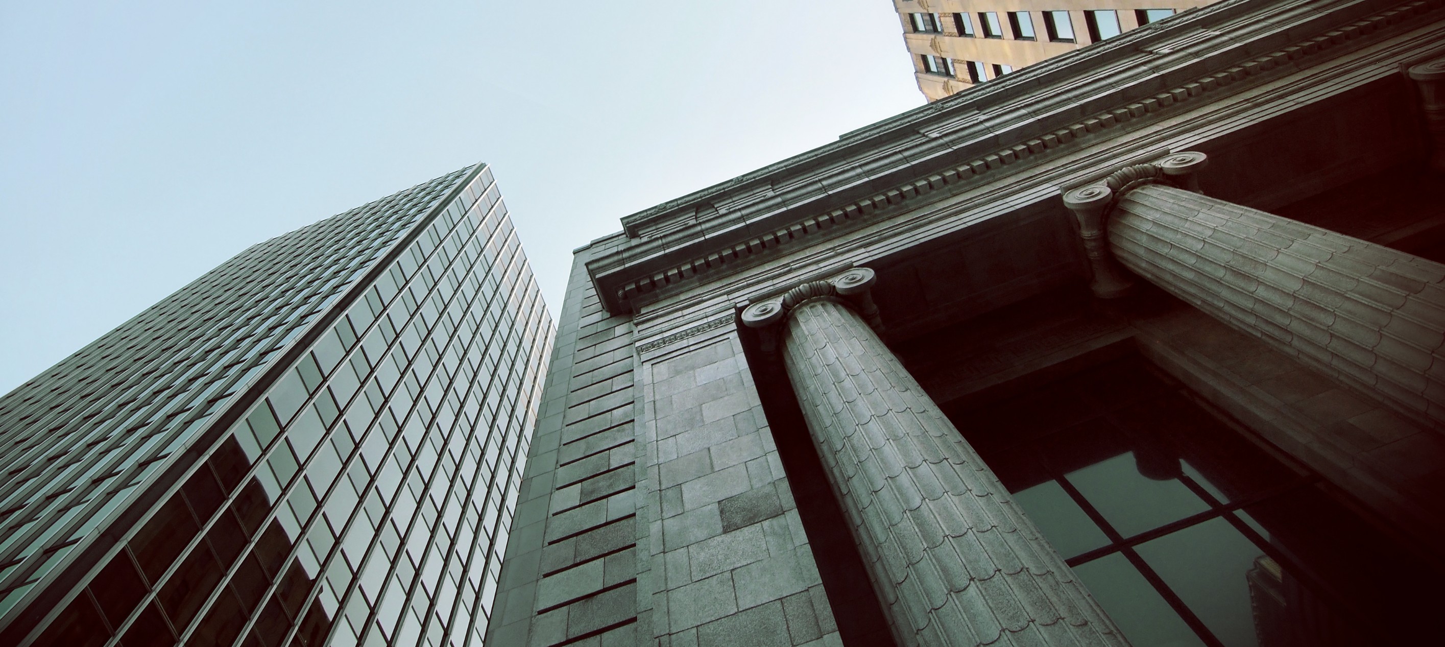 Looking up at a glass office tower beside a stone building with large fluted columns against a pale blue sky.