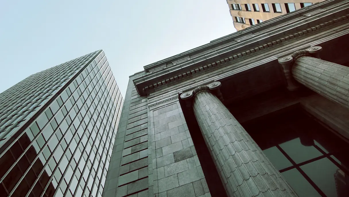 Looking up at a glass office tower beside a stone building with large fluted columns against a pale blue sky.