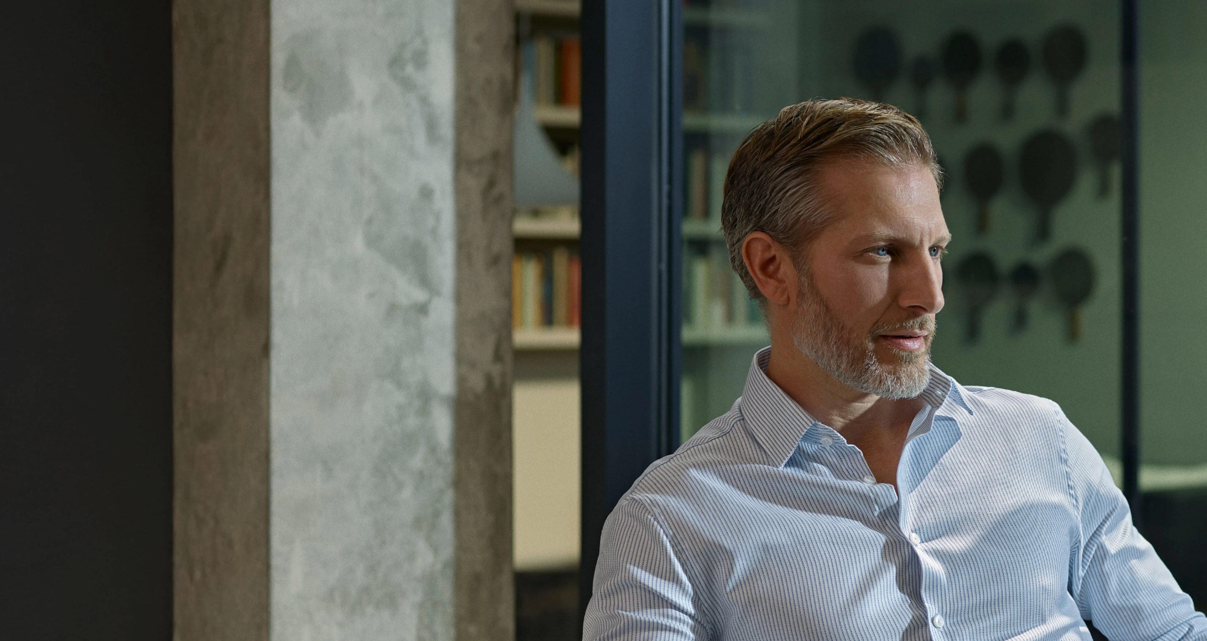 Man in a light blue striped dress shirt sitting indoors near a concrete column and bookshelf, holding a pen, with a dark glass panel behind him.