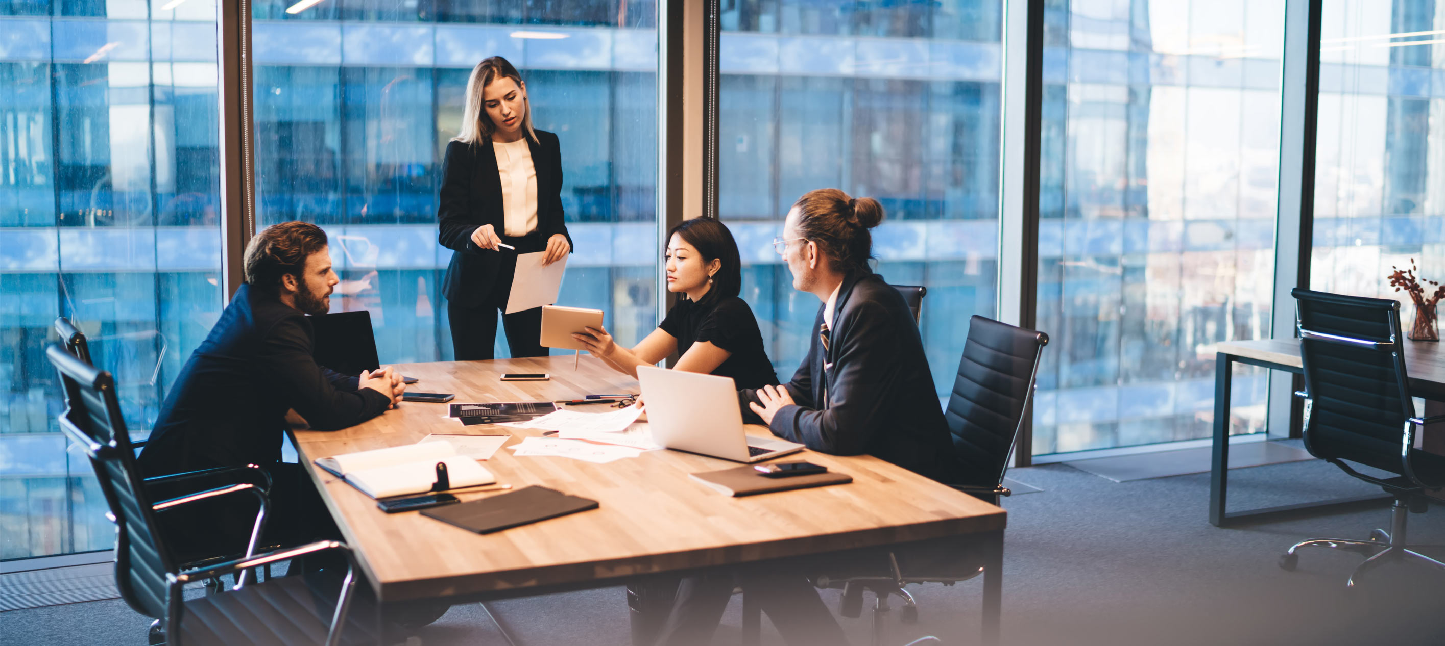 Four people in business attire sit and stand around a wooden meeting table with laptops, papers and a tablet.