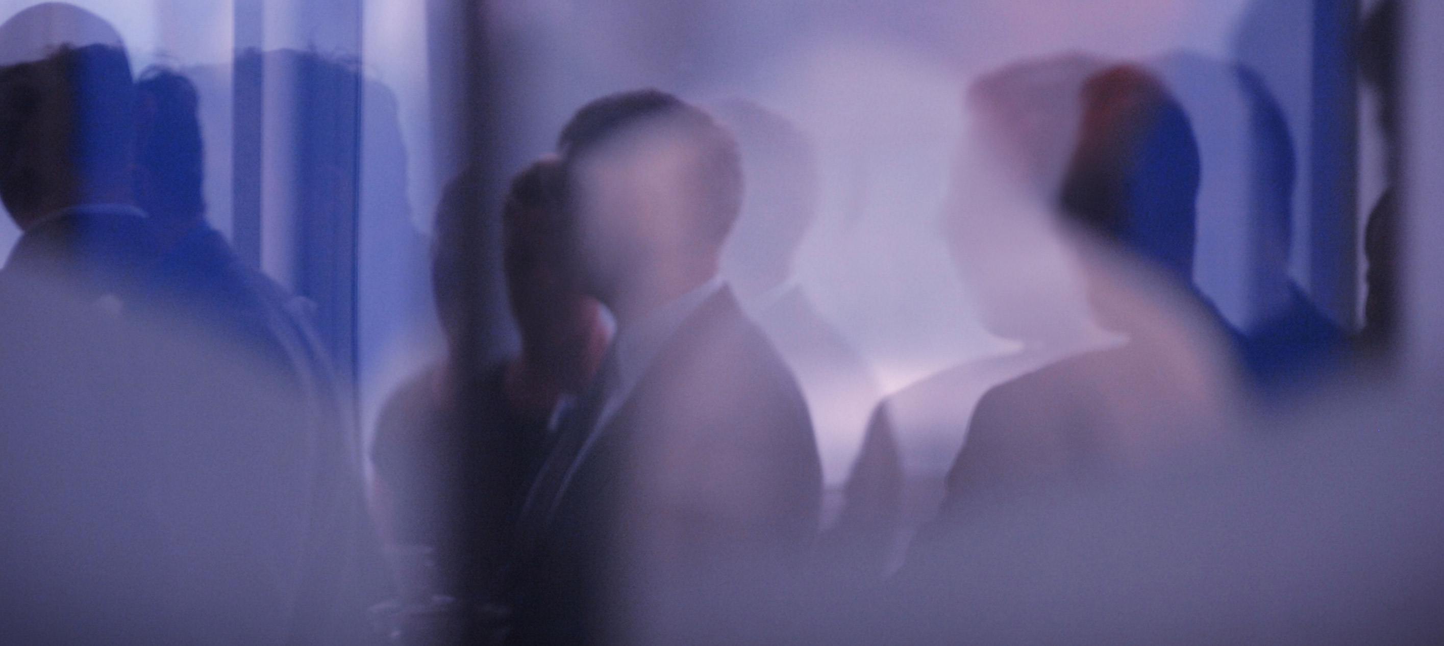 Blurry silhouettes of people standing in a room with soft blue and purple lighting, creating an indistinct group scene.