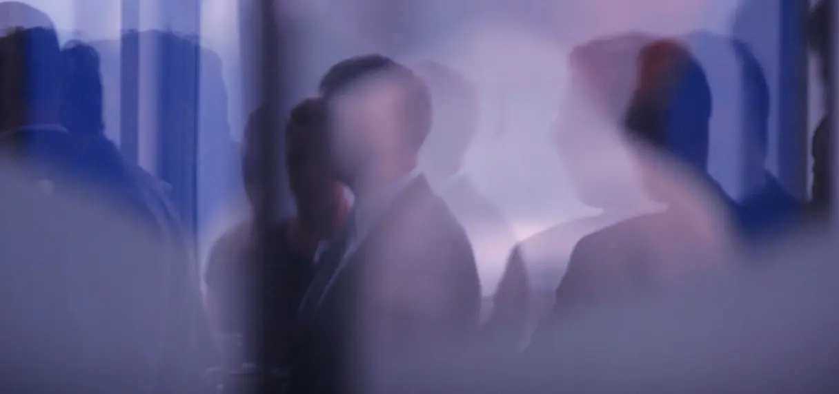 Blurry silhouettes of people standing in a room with soft blue and purple lighting, creating an indistinct group scene.