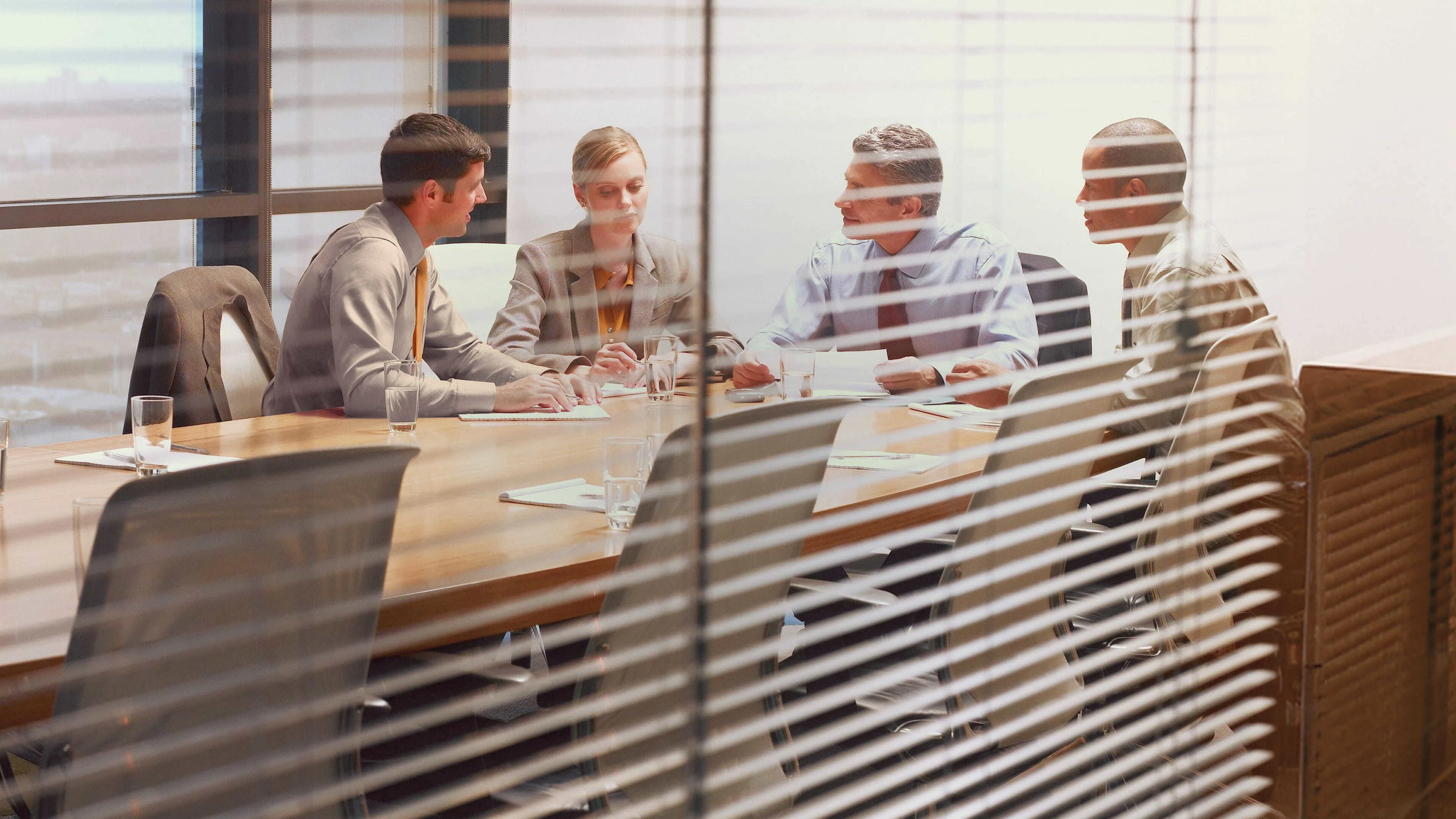 Four business professionals, three men and one woman, sit around a wooden conference table in a modern office, engaged in a serious discussion with documents and glasses of water.