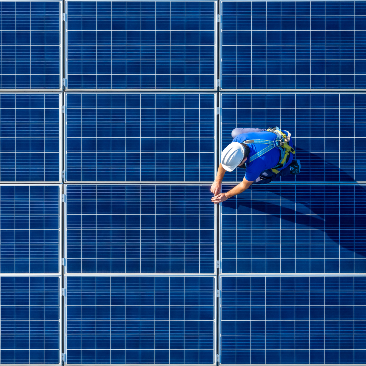 Engineer in a white helmet and safety harness working on a large array of blue solar panels, viewed from above, highlighting clean energy technology and sustainable practices.