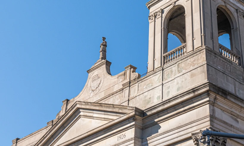 Stone church façade with bell tower topped by a cross, arched openings, statue on roofline and clear blue sky background.