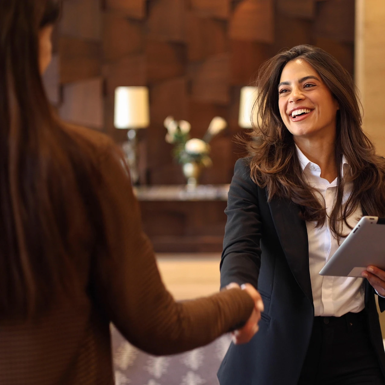 Two women shaking hands in a professional setting, one holding a tablet, with warm wooden walls and a lamp in the background.