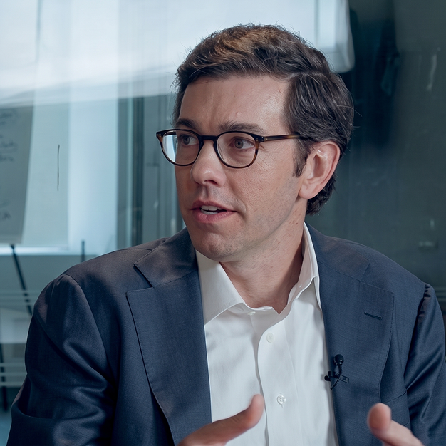 A man with brown hair and glasses wearing a dark suit and white shirt is speaking and gesturing in an office setting with glass walls.