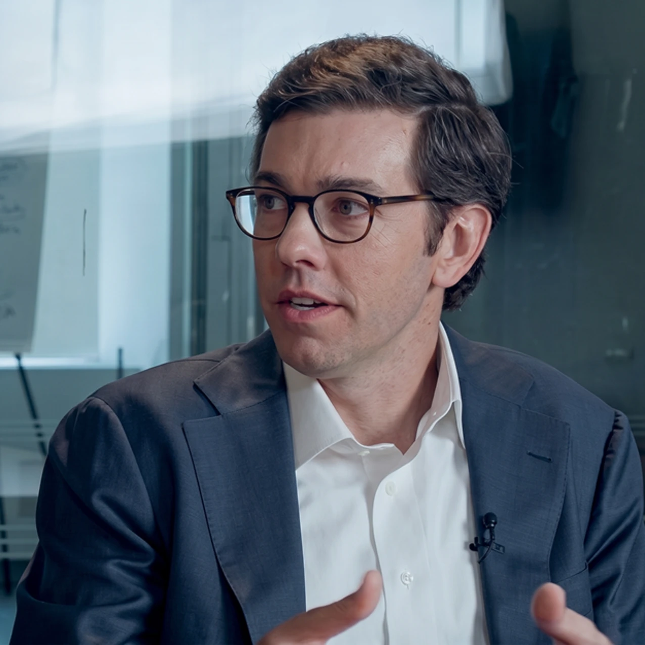 A man with brown hair and glasses wearing a dark suit and white shirt is speaking and gesturing in an office setting with glass walls.