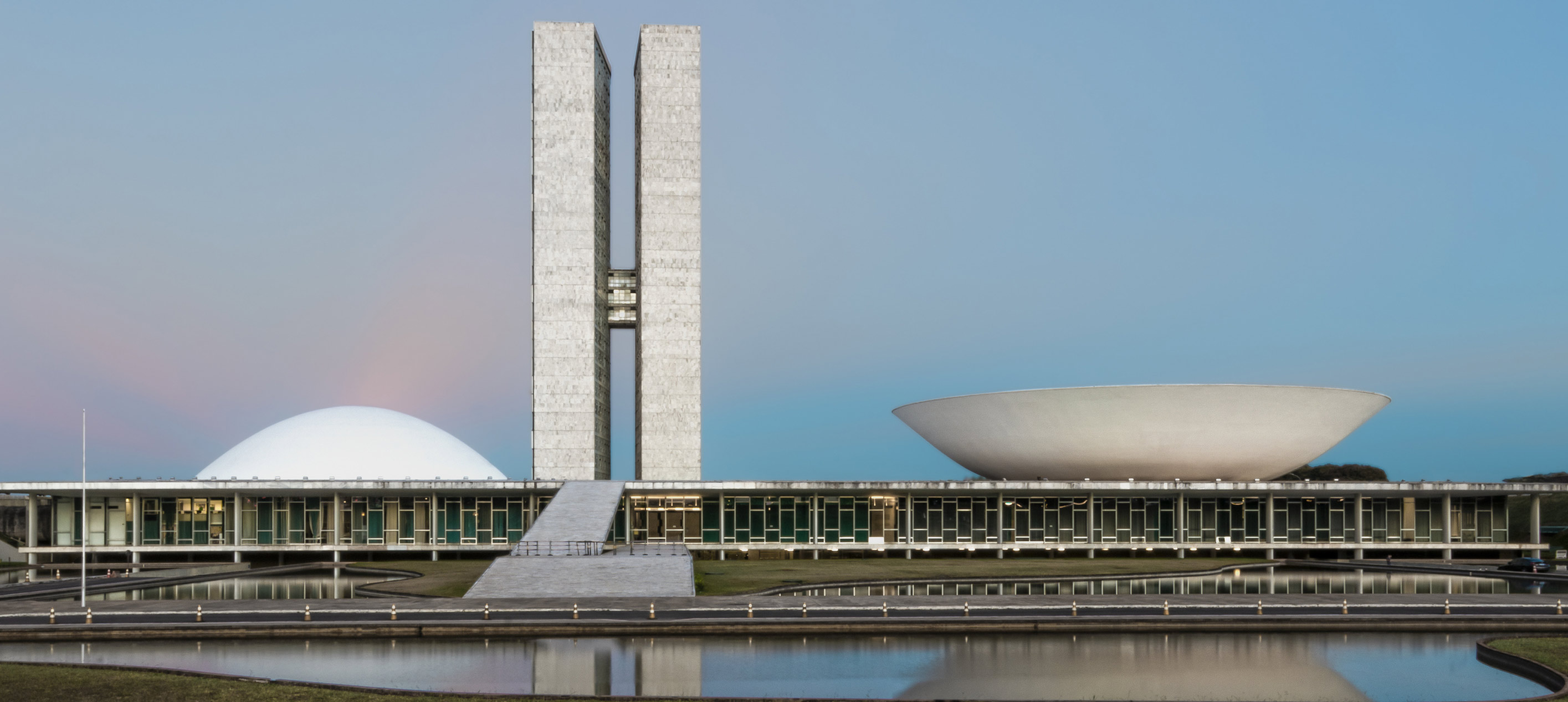 Two tall grey towers rise between a white dome and a white bowl-shaped roof on a long, low building by a pond.