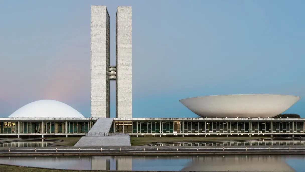 Two tall grey towers rise between a white dome and a white bowl-shaped roof on a long, low building by a pond.