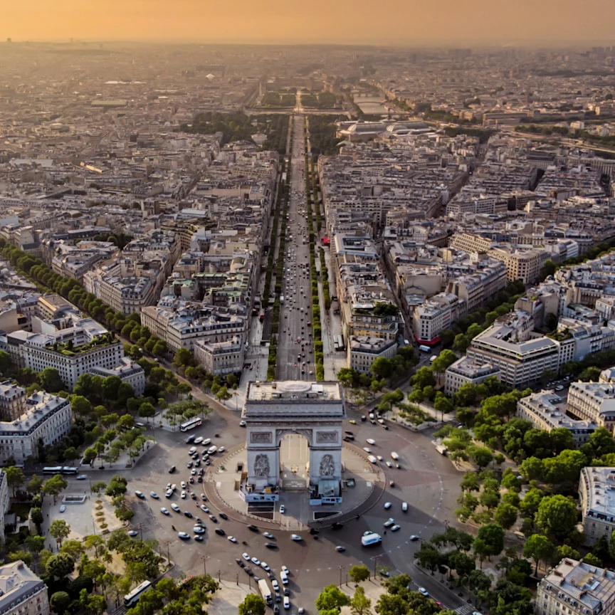 Aerial view of the Arc de Triomphe at sunset with busy circular traffic around it and long Parisian avenues extending into the city.