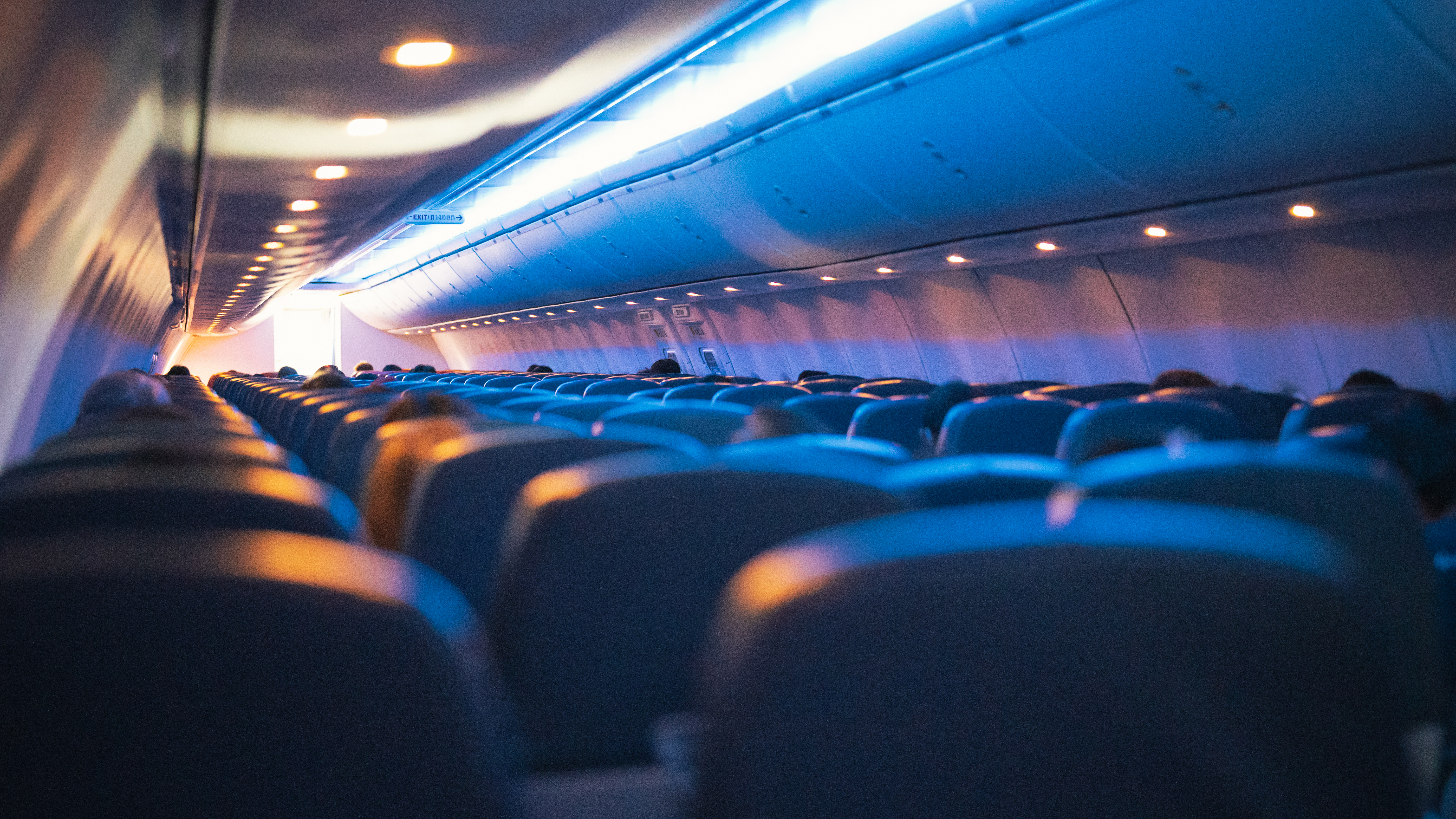 Rows of blue seats face the front of an aircraft cabin lit by blue and warm ceiling lights, with passengers seated.