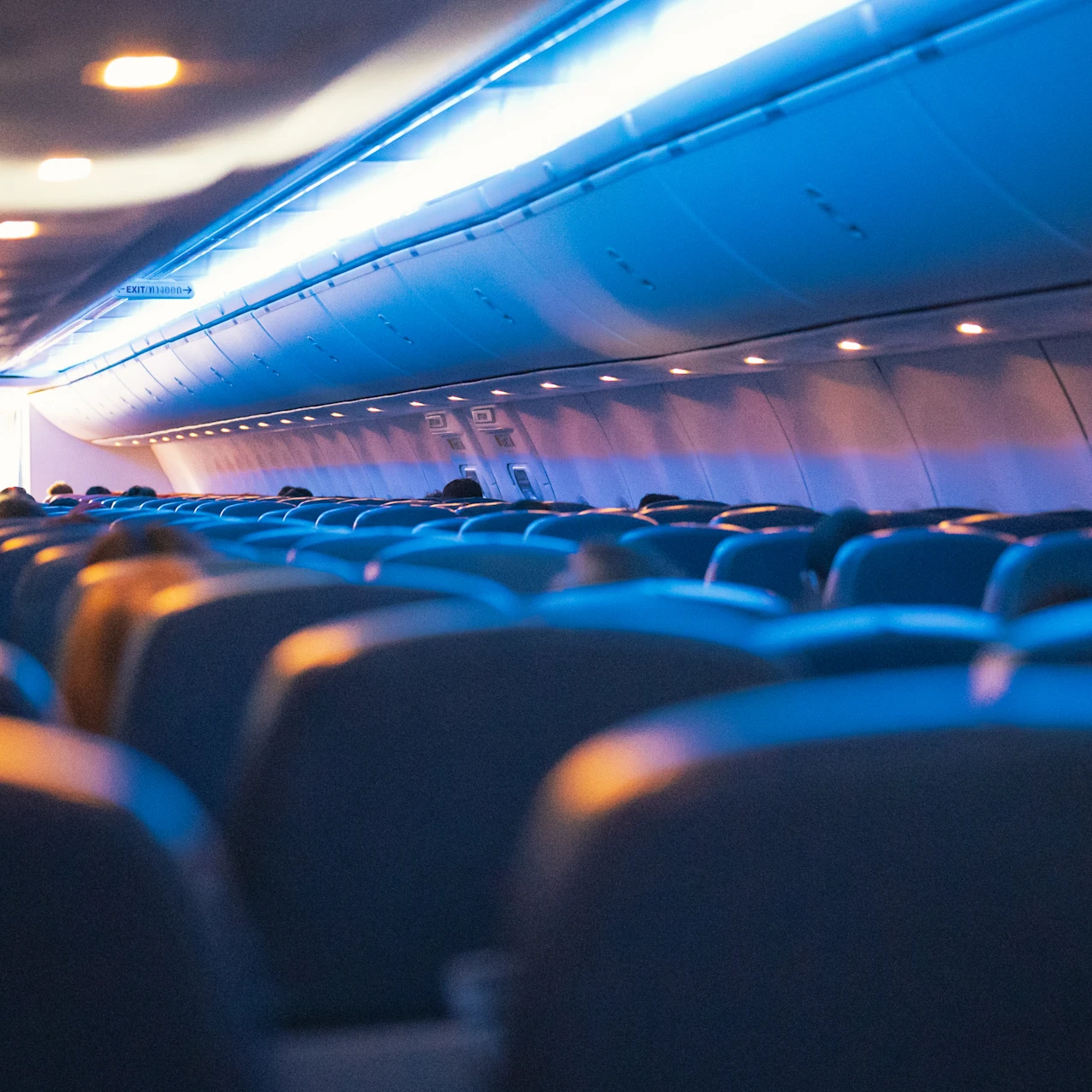 Rows of blue seats face the front of an aircraft cabin lit by blue and warm ceiling lights, with passengers seated.