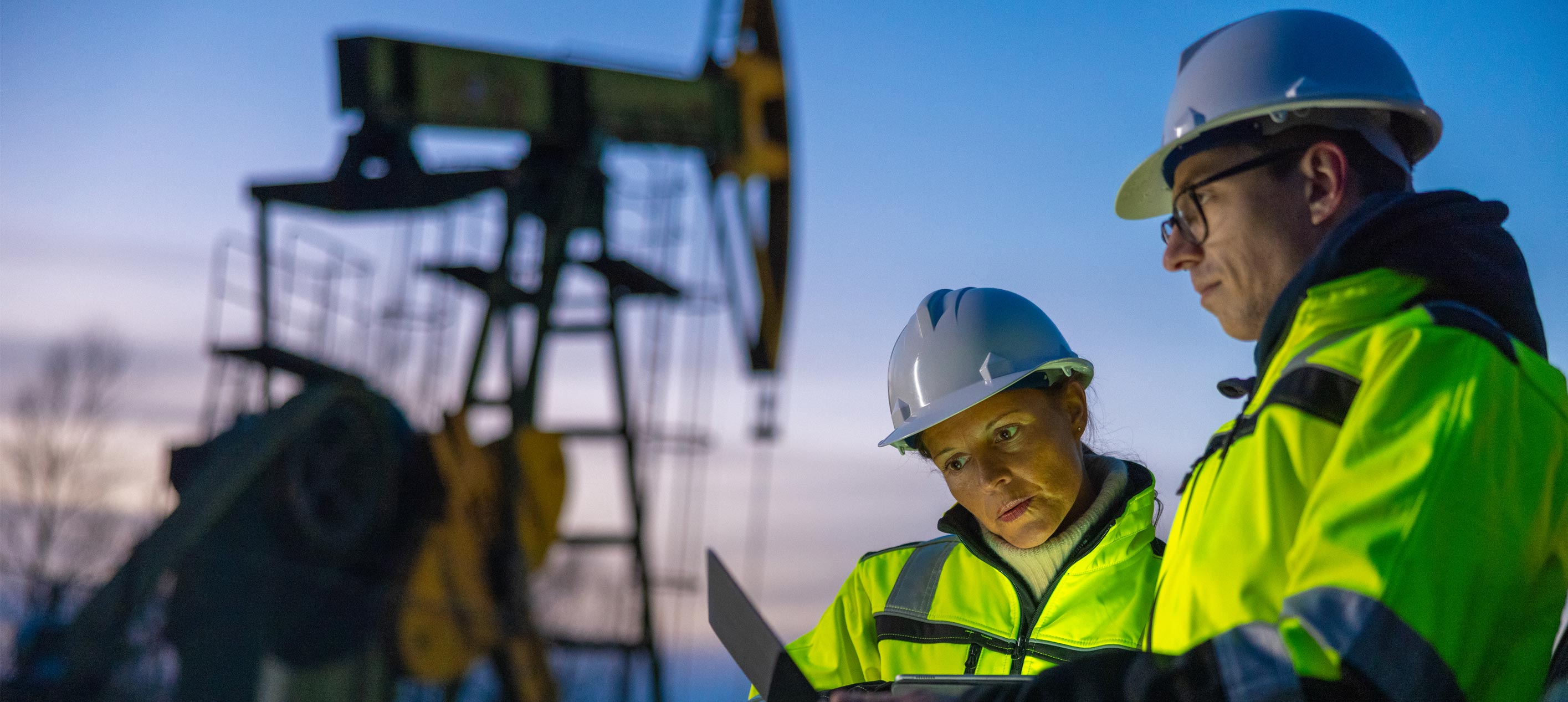 A man and woman in hard hats and hi-vis jackets look at a tablet in front of an oil pumpjack at dusk.
