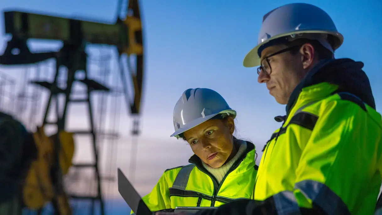 A man and woman in hard hats and hi-vis jackets look at a tablet in front of an oil pumpjack at dusk.