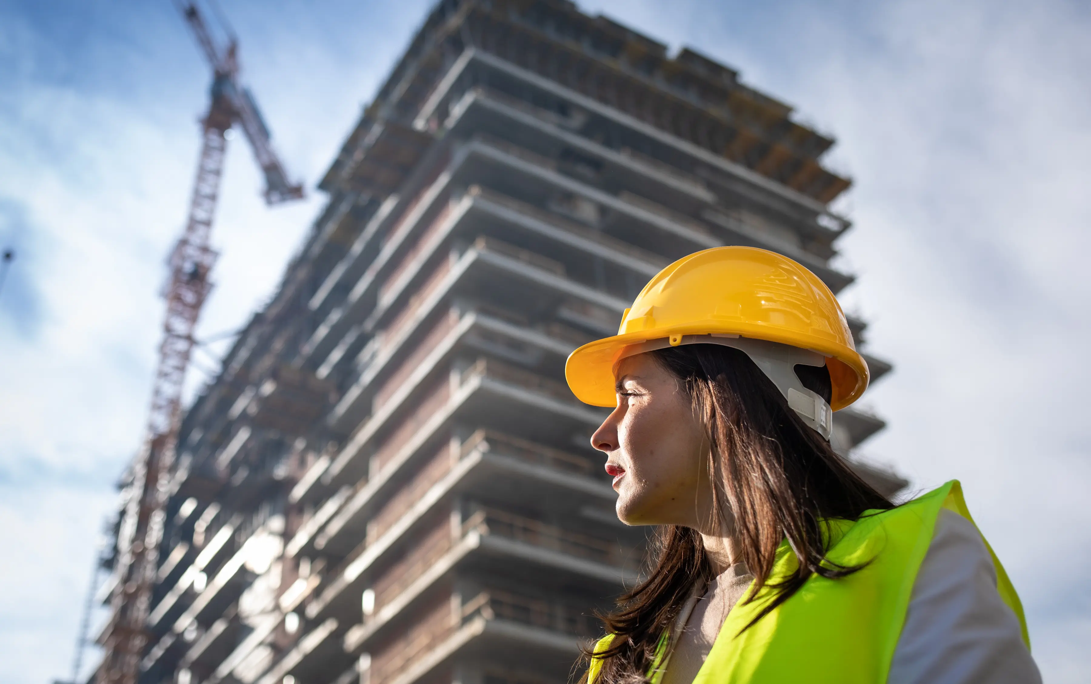 Construction worker wearing a yellow hard hat and reflective vest stands in front of a high-rise building under construction with a crane in the background.