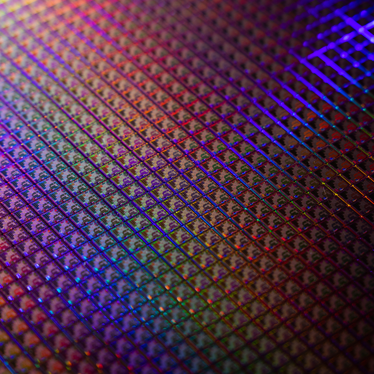 Close-up of a semiconductor wafer with intricate microchip patterns reflecting vibrant rainbow colors under bright light.